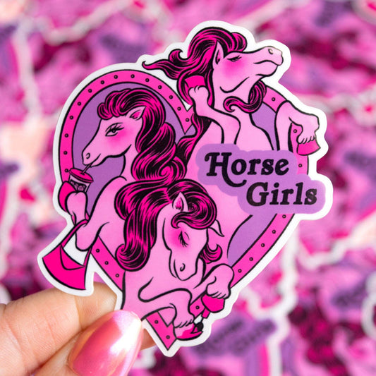A die-cut sticker featuring three stylized pink and purple horse girls, held by a hand with pink nail polish against a blurred pink patterned background. The sticker includes the text 'Horse Girls'.