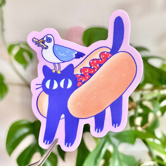 Sticker of a blue cat with a hot dog and seagull on a pink border against a green leafy background.
