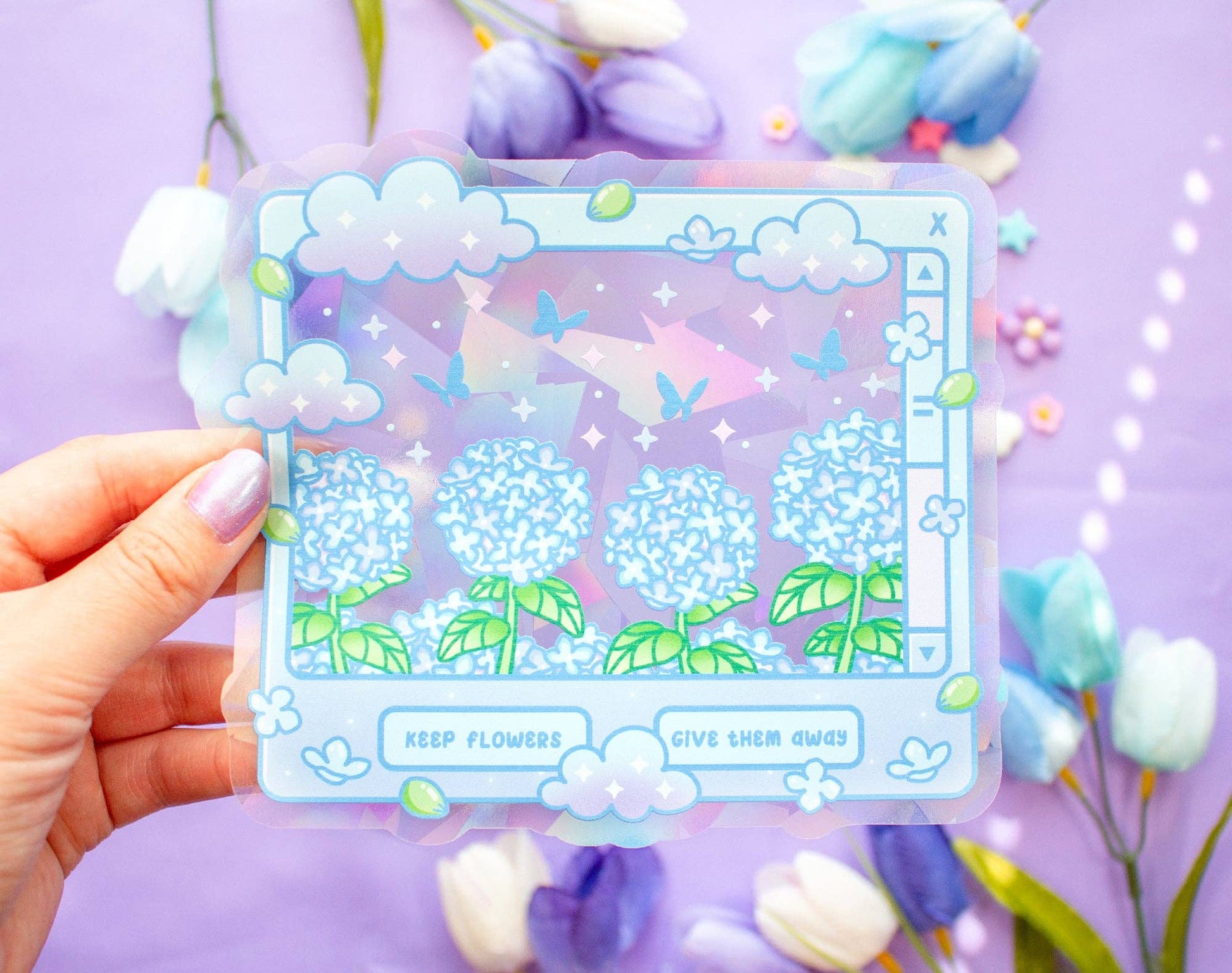 A hand holds a suncatcher featuring a pastel illustration of blue hydrangea-like flowers, butterflies, clouds, and the phrases 'KEEP FLOWERS' and 'GIVE THEM AWAY' on a lavender background with blurred flowers around it.