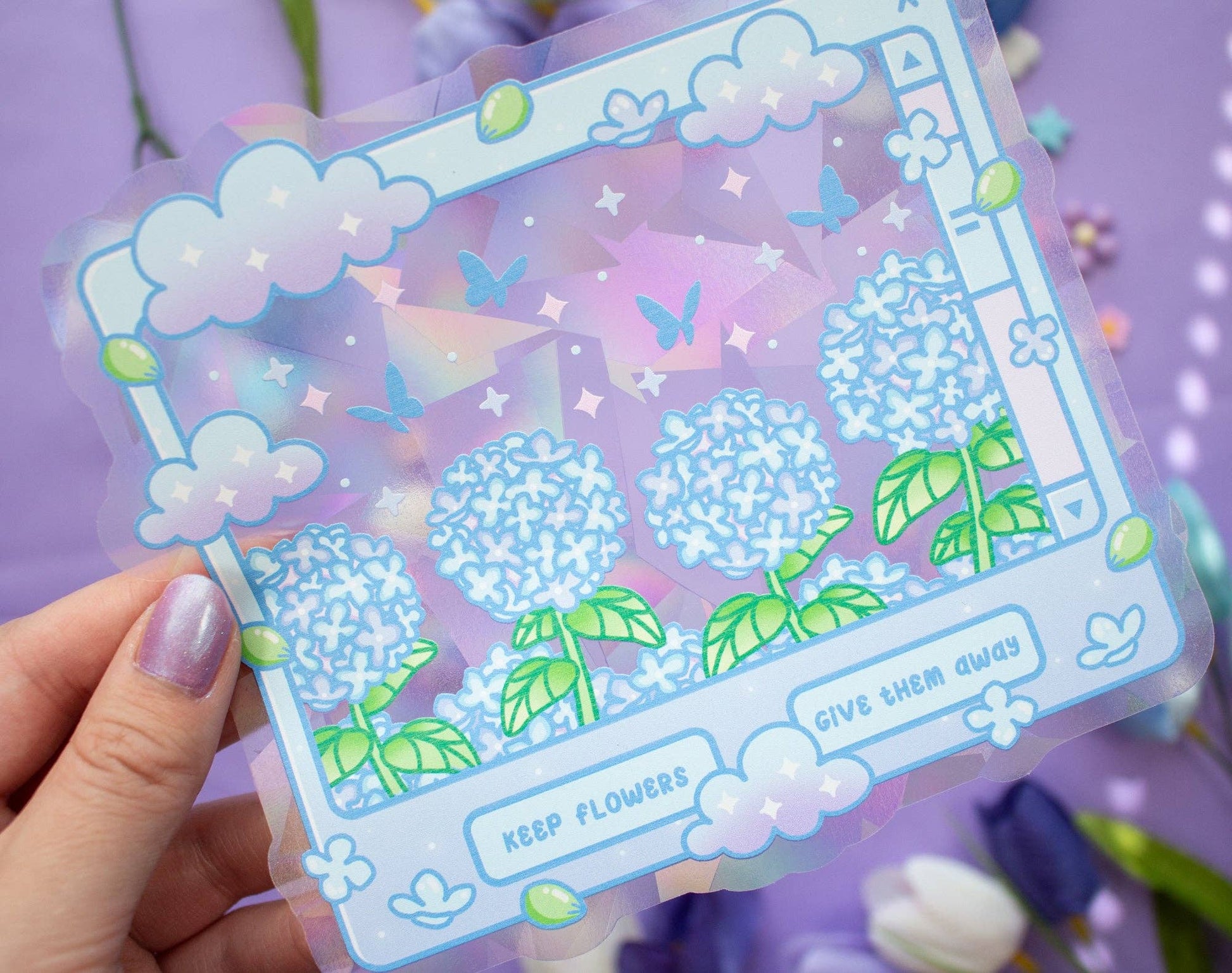 A hand with lavender nail polish holds a clear suncatcher sticker featuring a pastel illustration of blue hydrangea flowers, green leaves, clouds, butterflies, and decorative text on a purple background.