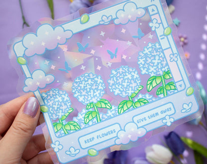 A hand with lavender nail polish holds a clear suncatcher sticker featuring a pastel illustration of blue hydrangea flowers, green leaves, clouds, butterflies, and decorative text on a purple background.