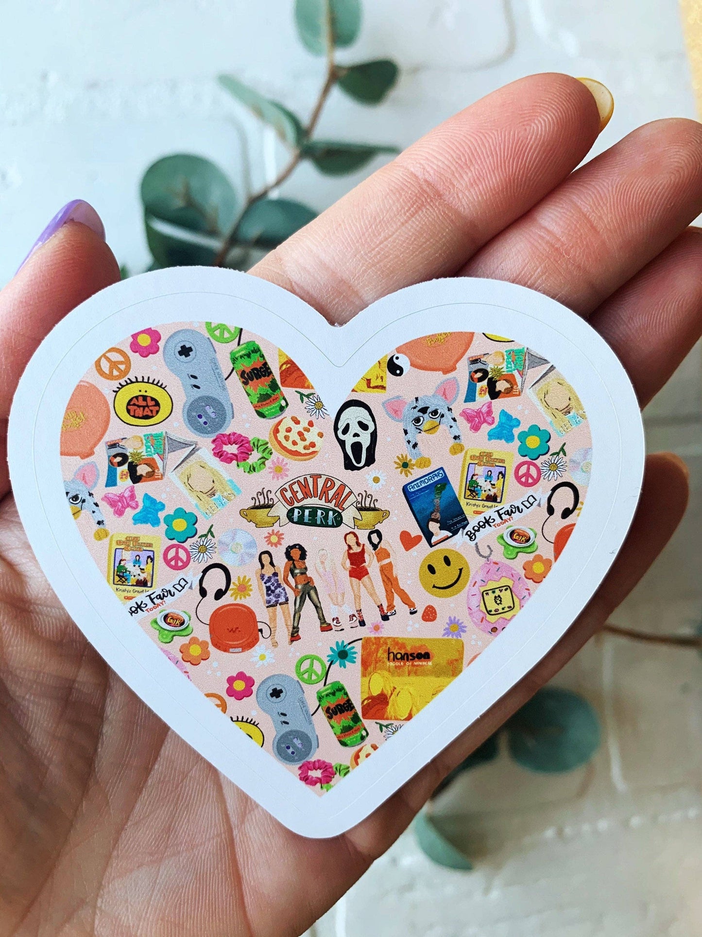 A heart-shaped sticker held in a person's hand. The sticker features a white border and a colorful collage of various pop culture icons, characters, and phrases inside.