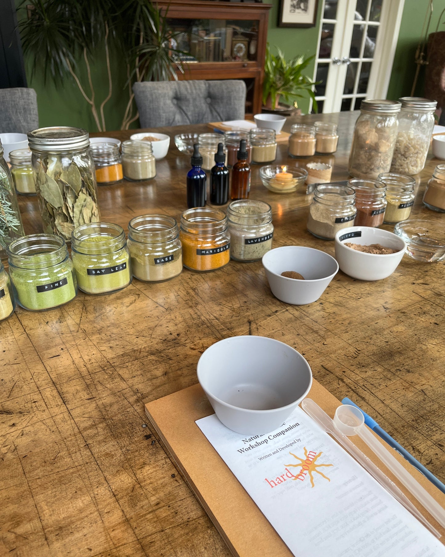 A tabletop display of handmade herbal products including glass jars filled with dried herbs, small white bowls, dark glass bottles, and a printed recipe card on a wooden board, set in a rustic indoor room with a wooden table and plants in the background.