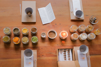 An overhead view of a wooden table arranged with small glass jars containing colorful powders, white cups, printed cards, and an open box with packaged items. No people or animals are present, but this is not a clean isolated product shot; it shows the products arranged in a real tabletop setting, so Lifestyle fits better than Product/Packaging.