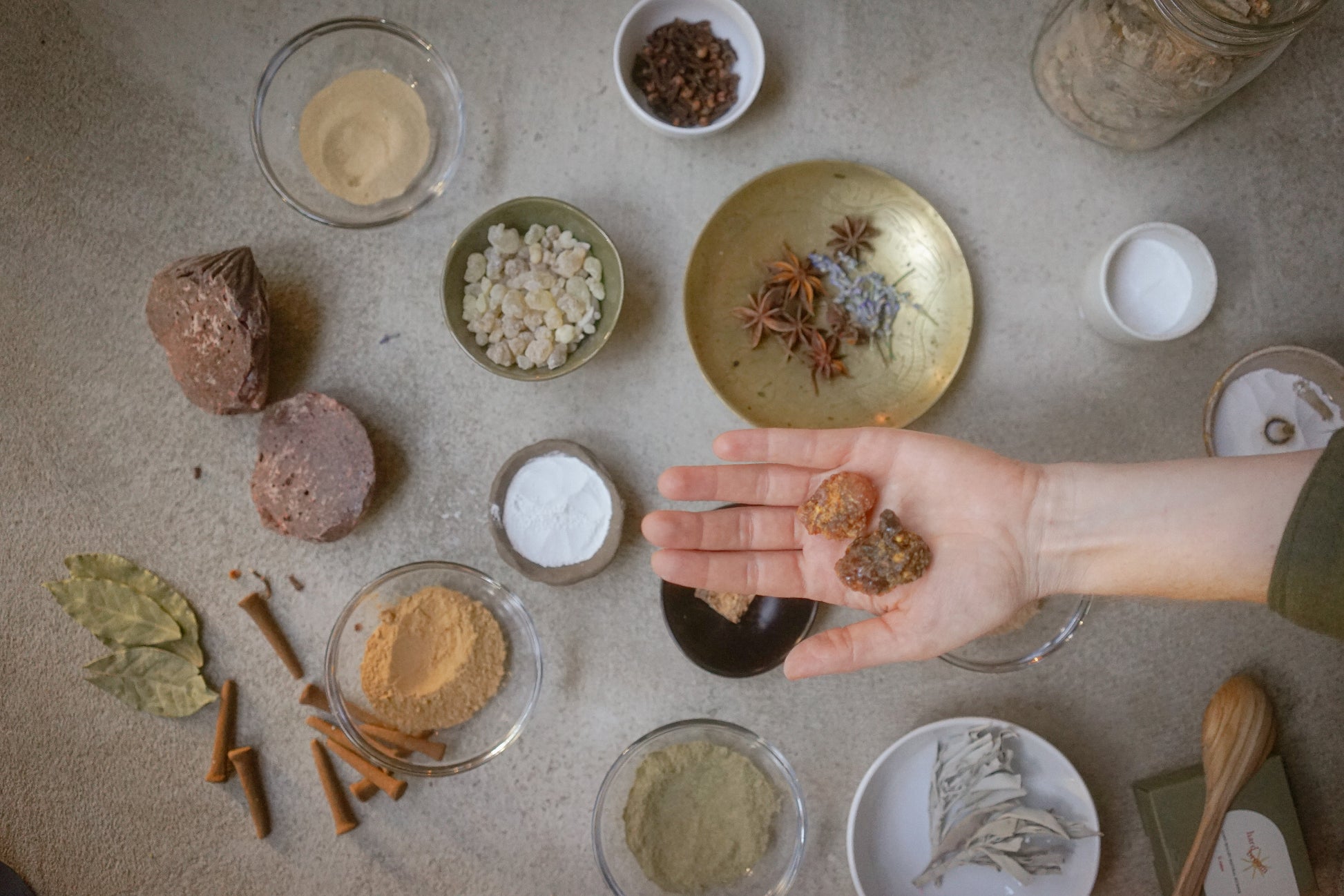 An overhead flat-lay of various natural ingredients and spices arranged on a textured gray surface, with a hand holding two dried botanical pieces in the center.