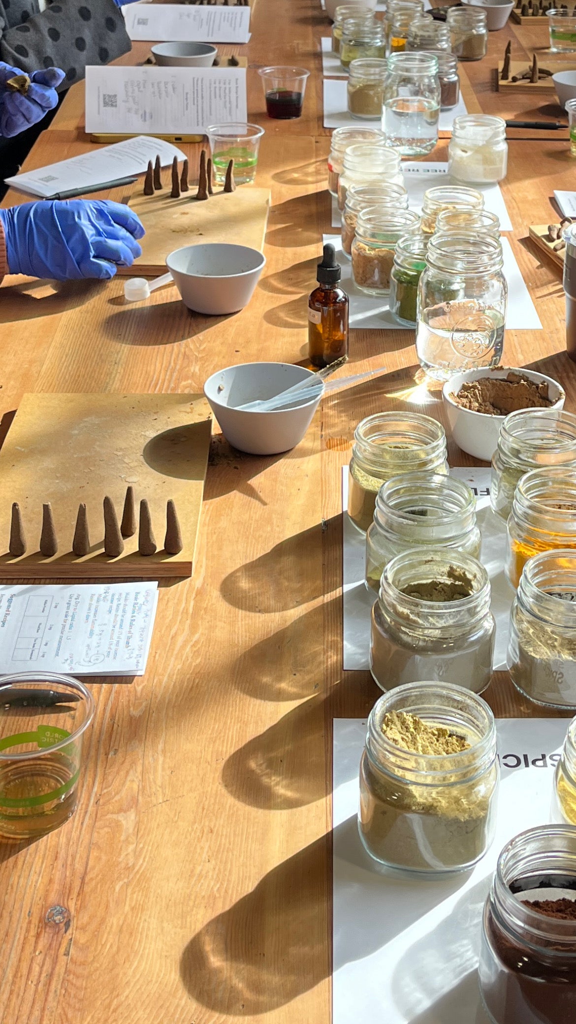 A wooden table set up with multiple glass jars containing various powders and herbs, white bowls, small bottles, and a person wearing blue gloves reaching in. Papers and tools are arranged along the table in a workshop or laboratory setting.