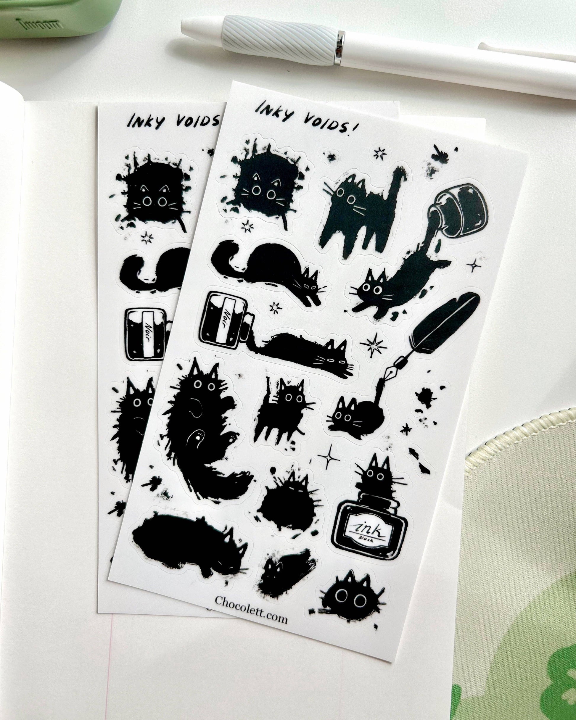 Two sheets of black inkblot-style stickers on a white surface with a pen and green object in the background.