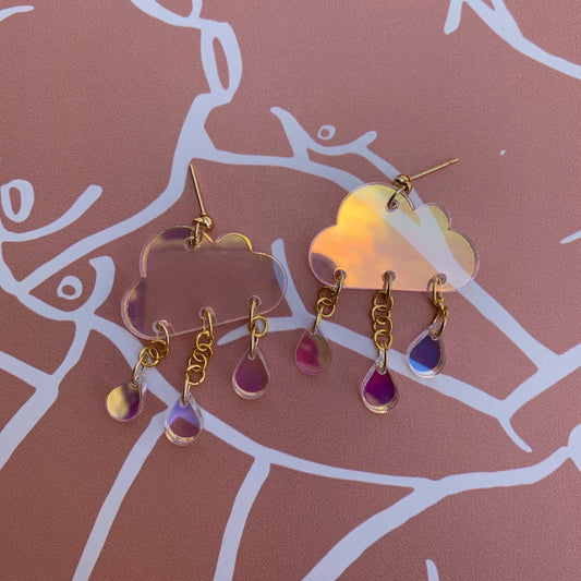 Pair of cloud-shaped earrings with gold chains and purple drops on a pink background.