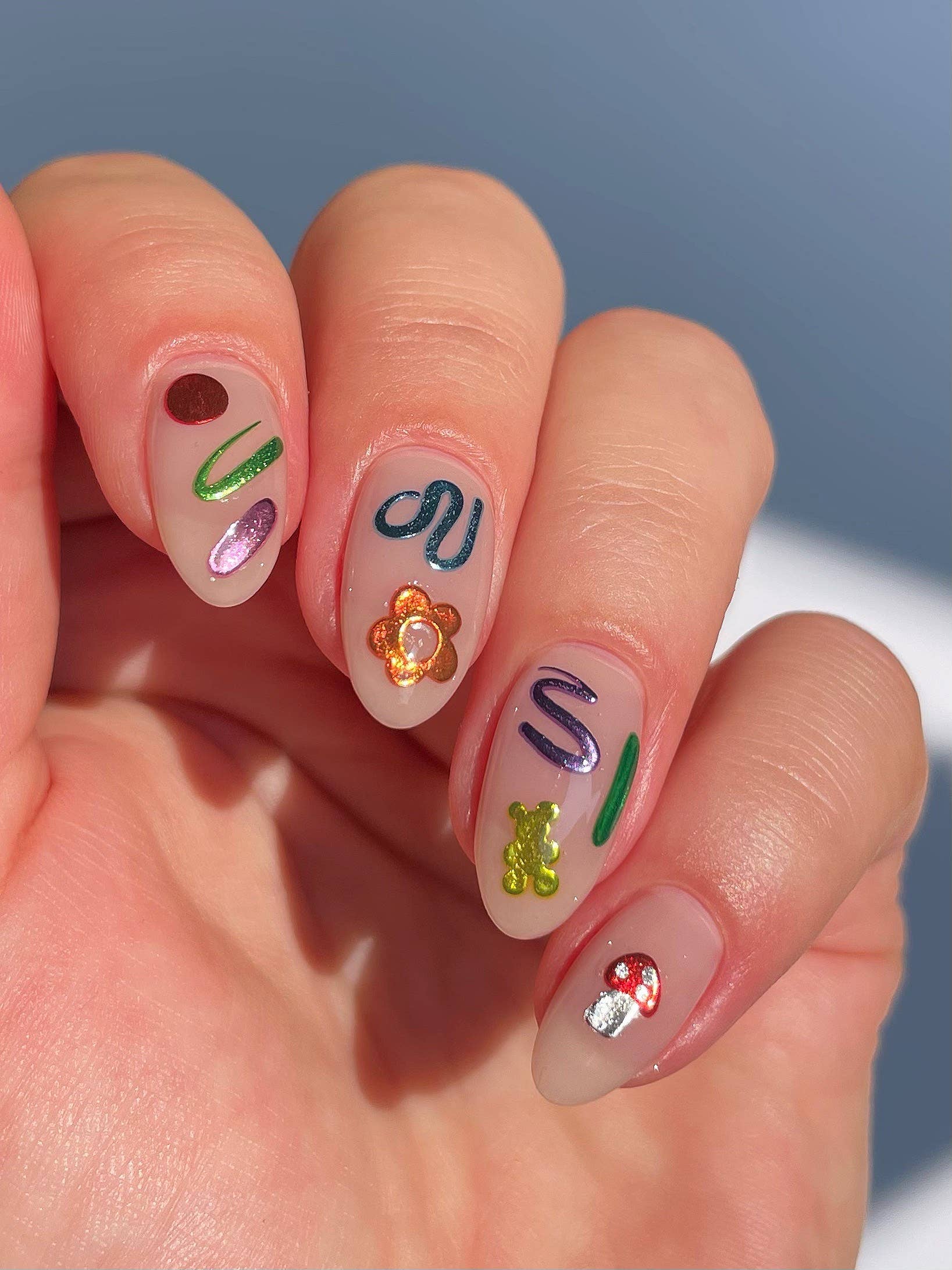 A close-up of a hand with manicured nails decorated with colorful, whimsical nail art including abstract shapes, a flower, a gummy bear, and a mushroom on a pale pink base.