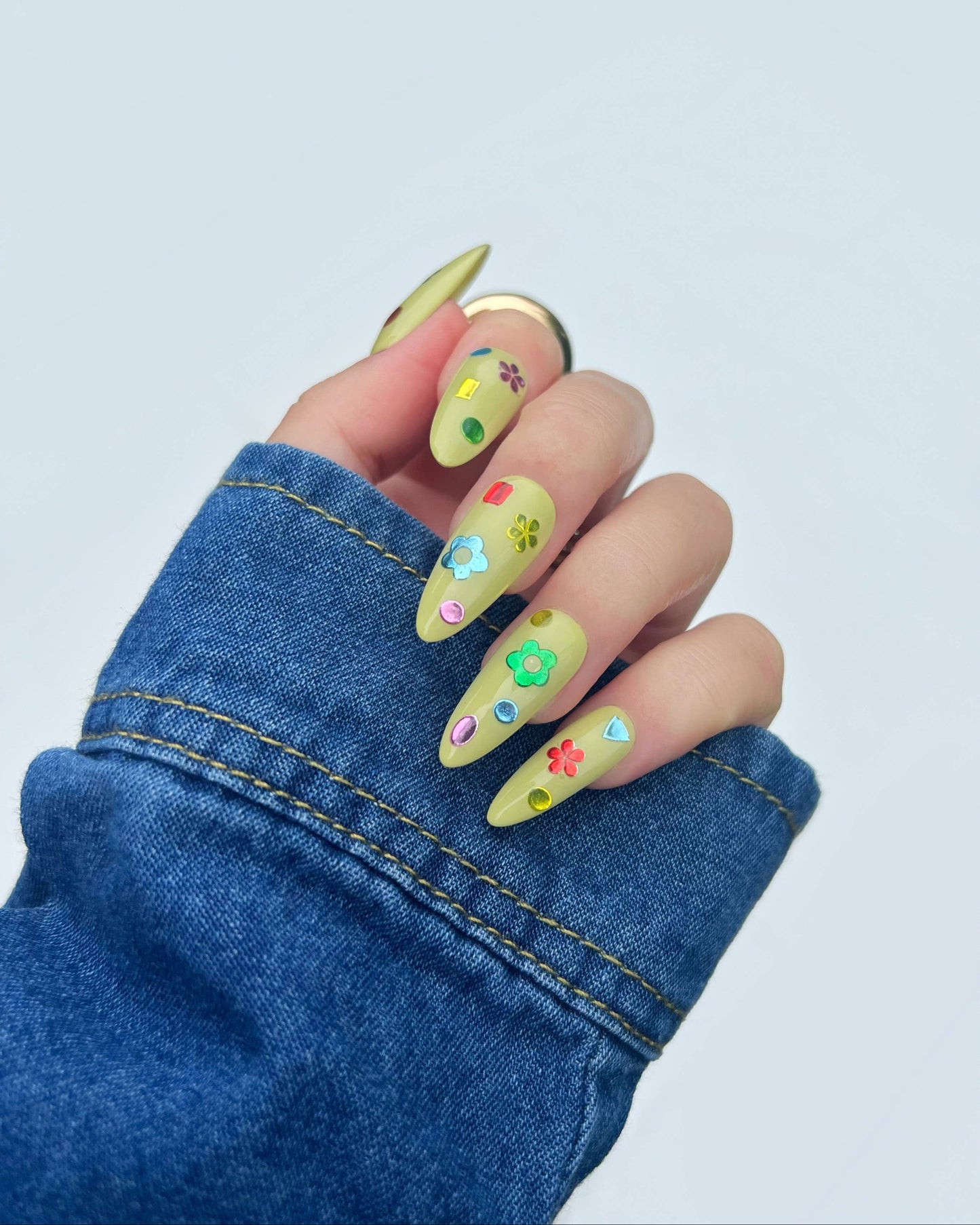 A close-up of a hand with long almond-shaped nails painted in a light yellow-green base color and decorated with small colorful floral and heart motifs. The hand is wearing a blue denim sleeve against a clean, light gray background.