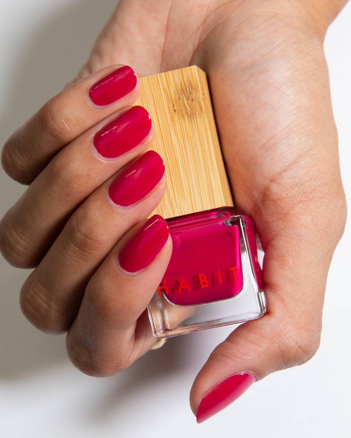 Hand holding a red nail polish bottle with a wooden cap on a light background