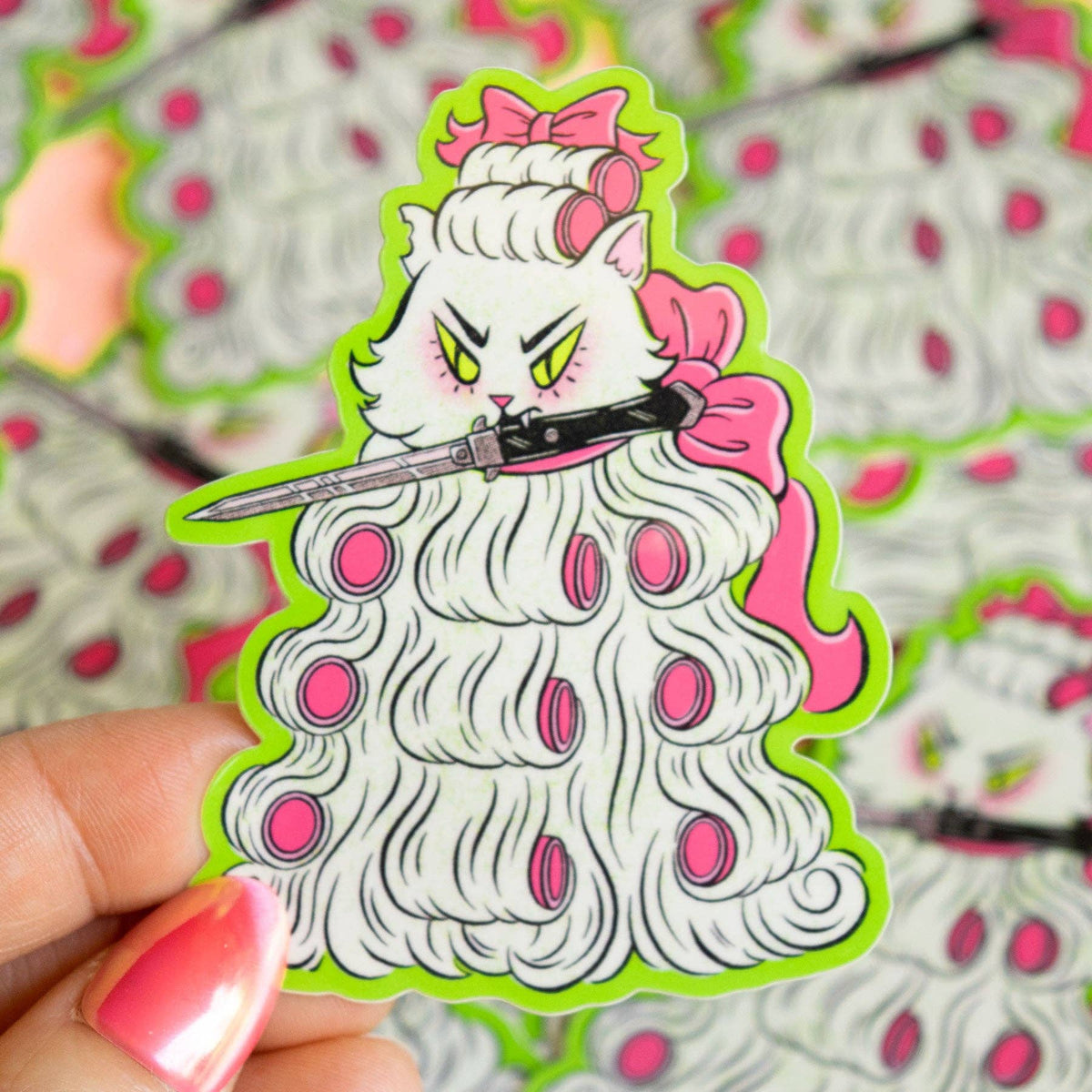 A hand holds a die-cut sticker featuring an angry white cat with pink hair accessories, yellow eyes, and a knife, set against a background of multiple identical stickers.
