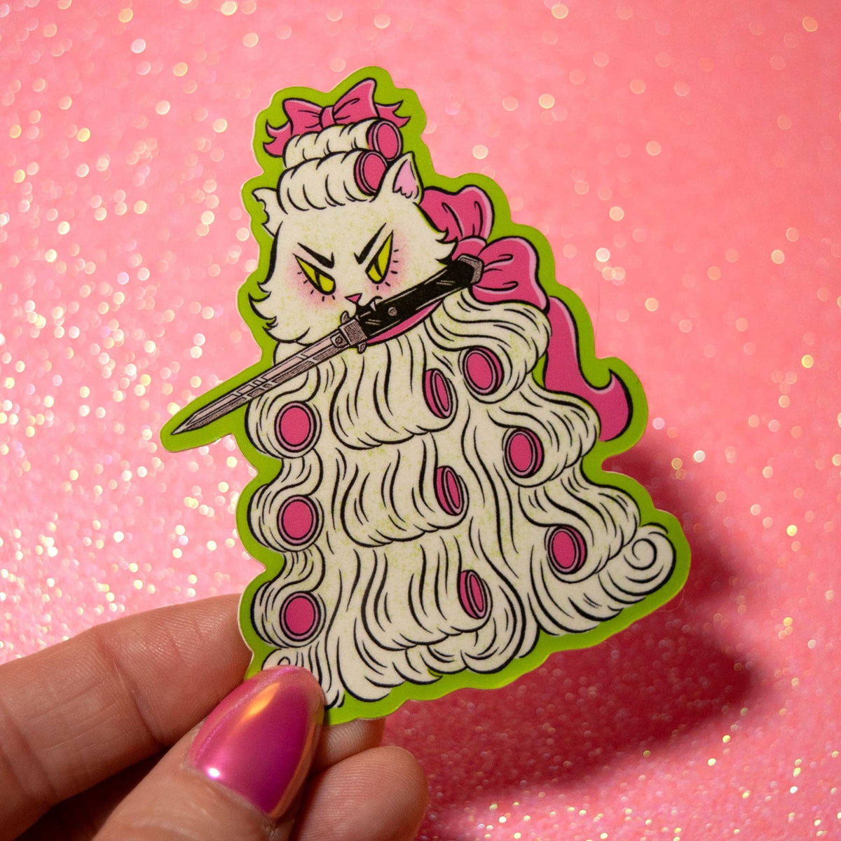 A hand with pink manicured nails holds a die-cut sticker or pin featuring a stylized cat with long white hair, pink bows, and a sword, set against a pink glitter background.