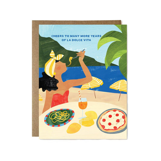Greeting card with illustration of a woman at an outdoor table with food and drink, with text 'Cheers to many more years of La Dolce Vita'.