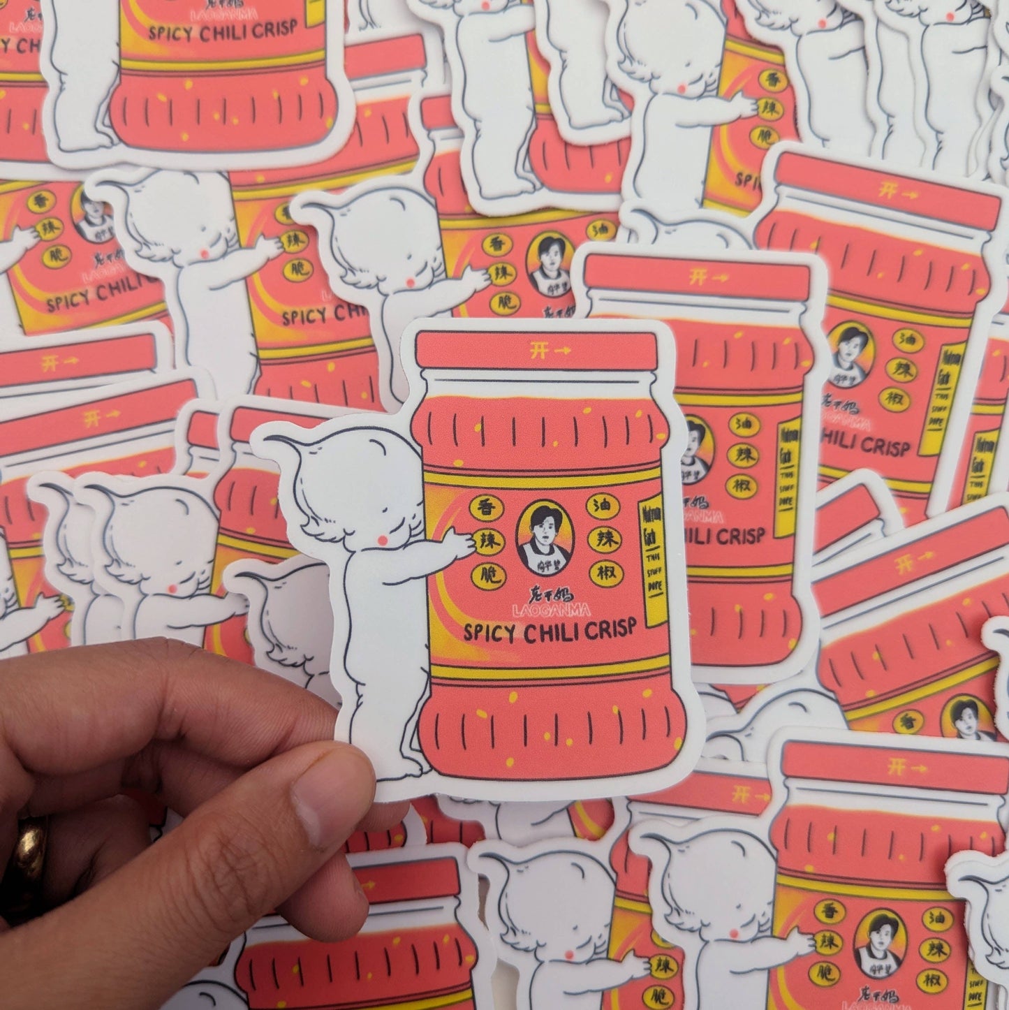Sticker of a red container labeled 'Spicy Chili Crisp' held over a patterned background with similar designs. Lao Gan Ma + Kewpie Vinyl Sticker