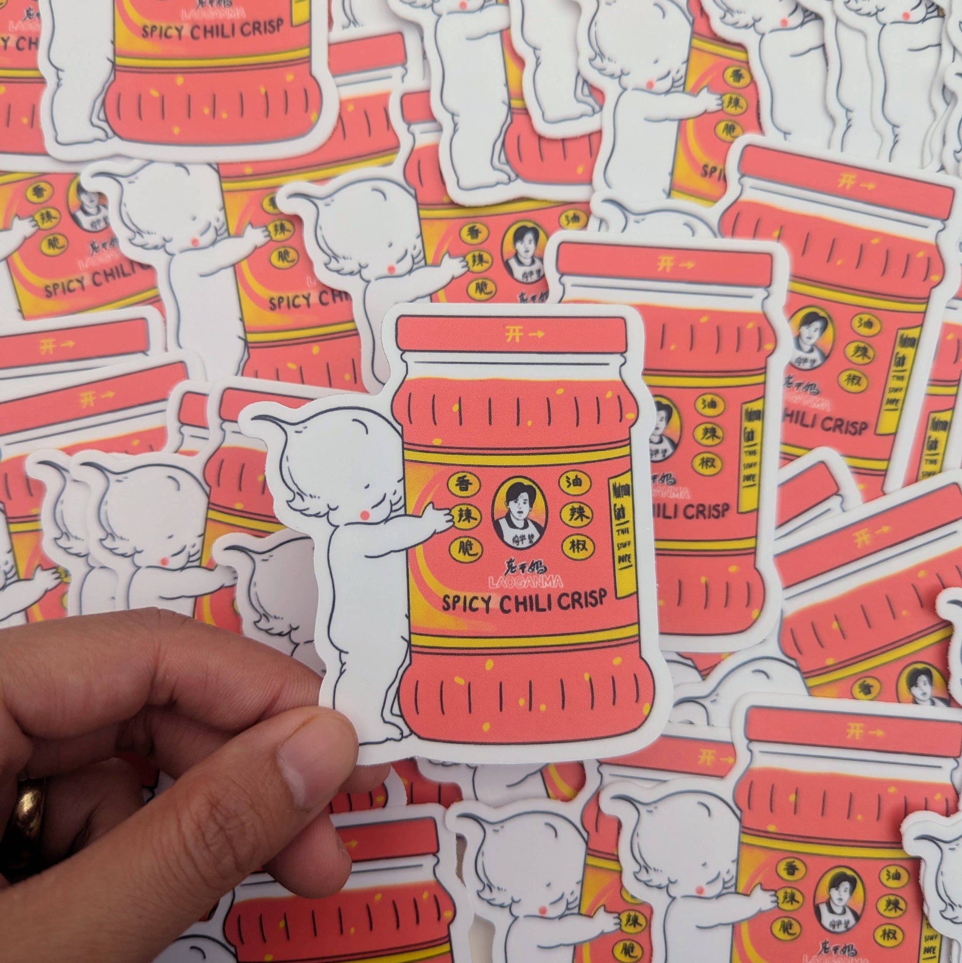 Sticker of a red container labeled 'Spicy Chili Crisp' held over a patterned background with similar designs. Lao Gan Ma + Kewpie Vinyl Sticker