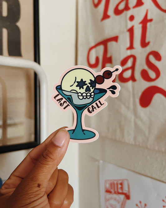 A hand holds a die-cut sticker featuring a cartoon skull in a teal cocktail glass with the words 'LAST CALL' , set against a background of a white wall with a canvas print and a lamp.