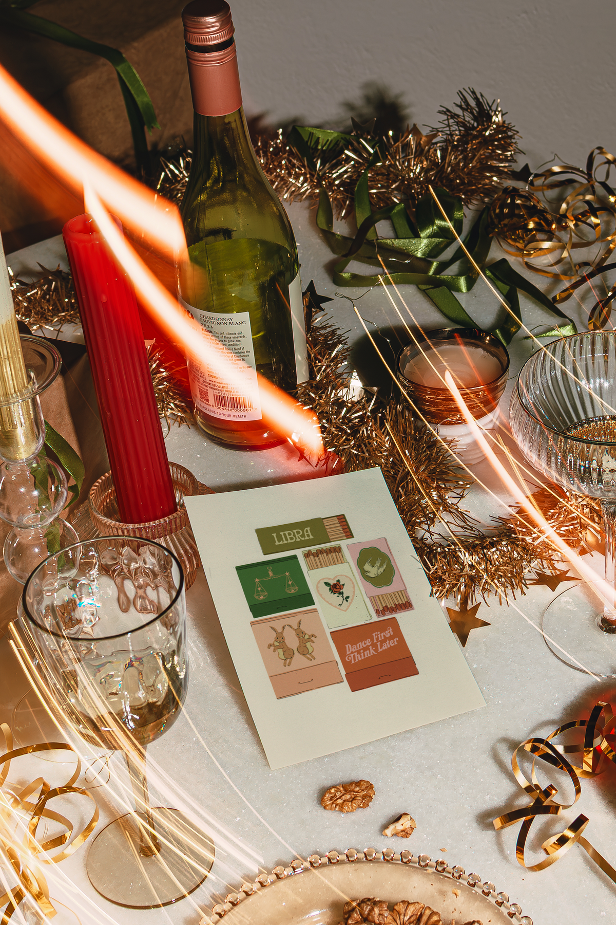 A styled holiday tabletop scene with a printed card or booklet in the center, surrounded by a green wine bottle, red candle, gold tinsel, sparklers, glasses, and nuts on a table. No person is visible, but this is not a clean isolated product shot because a real environment and surrounding decor are present, so subject_type is Lifestyle.