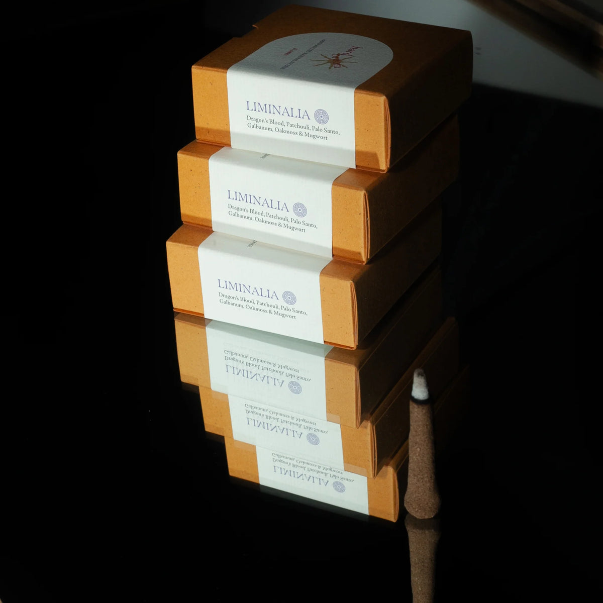 Three stacked Liminalia incense boxes in warm orange-brown packaging with white paper bands, photographed on a glossy black reflective surface.