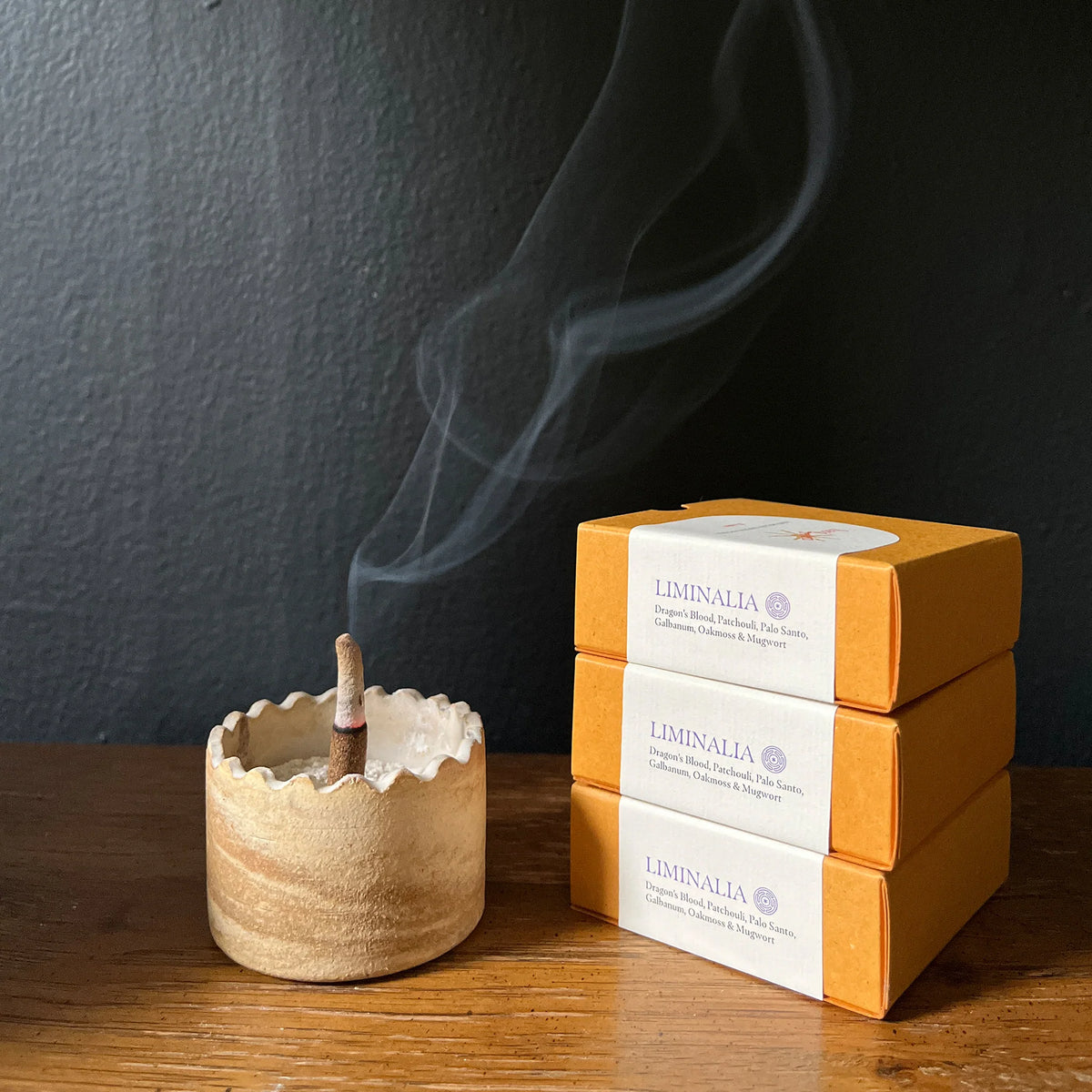 Three orange boxes of Liminalia incense sticks with white paper bands are stacked next to a small ceramic incense holder with a smoking stick, set on a wooden table against a dark gray wall.