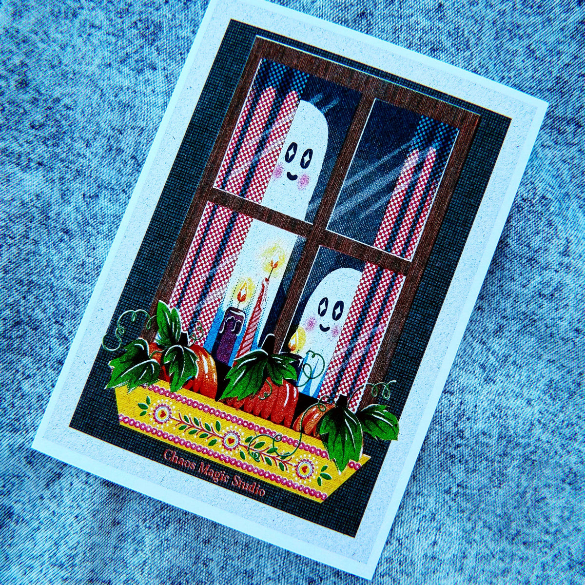 A square printed card or art print featuring a whimsical Halloween scene: two white ghosts peeking through a window with red-and-white striped curtains, candles, and a flowered yellow ledge with red apples. The card has a wide white border and is photographed on a blue textured fabric background.