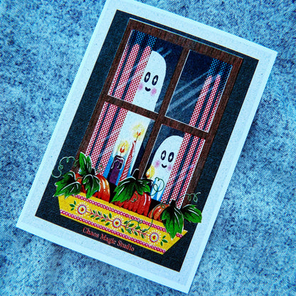 A square printed card or art print featuring a whimsical Halloween scene: two white ghosts peeking through a window with red-and-white striped curtains, candles, and a flowered yellow ledge with red apples. The card has a wide white border and is photographed on a blue textured fabric background.
