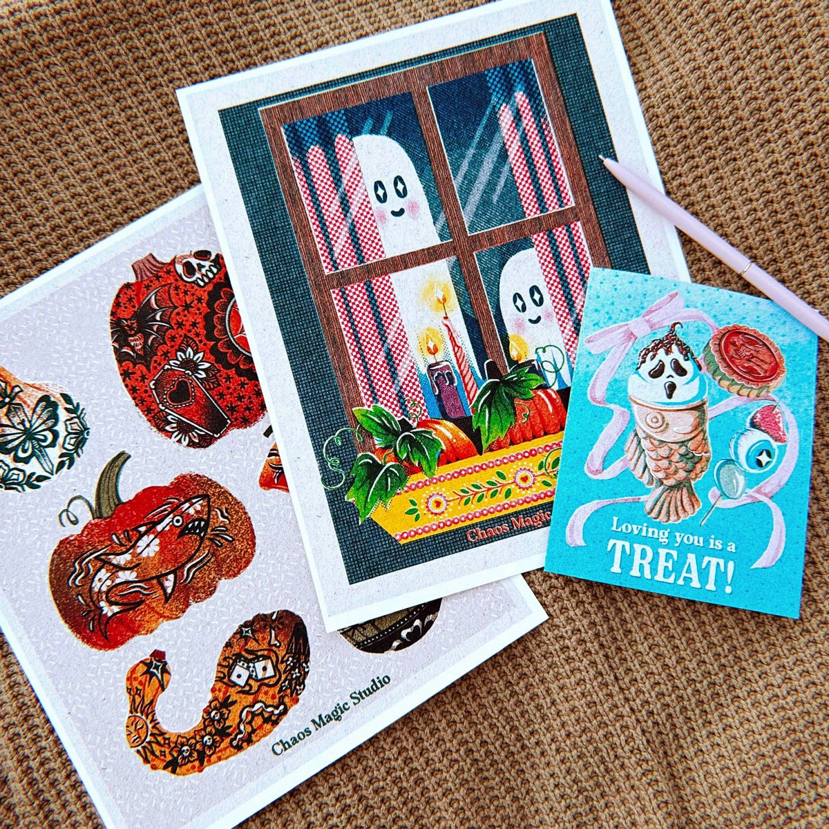 A styled flat-lay of two Halloween-themed art prints on a brown woven fabric background. The larger print shows cute cartoon ghosts looking out a window with candles and pumpkins, and the smaller print says 'Loving you is a TREAT!' with a ghost and candy cane.