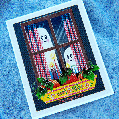 A square printed card or art print featuring a whimsical Halloween scene with two white ghosts behind a window, red-and-white striped curtains, candles, pumpkins, and a yellow flowered ledge. The card is photographed on a textured blue fabric background.