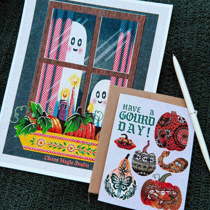Two illustrated greeting cards laid on a dark textured surface. The larger card shows a window scene with ghost figures, candles, pumpkins, and the text 'Chaos Magic Studio.