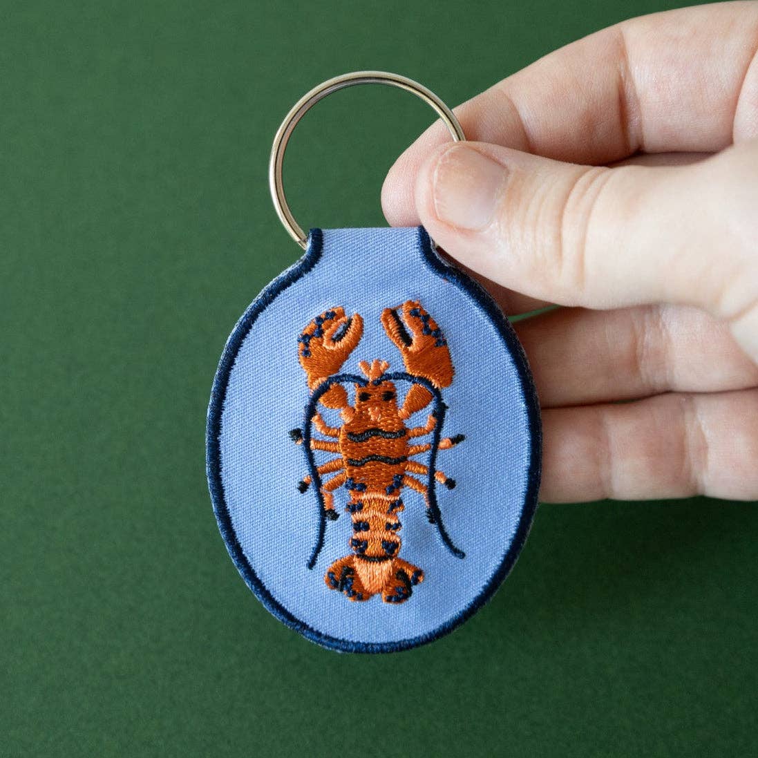 Keychain with embroidered lobster design held by a hand against a green background
