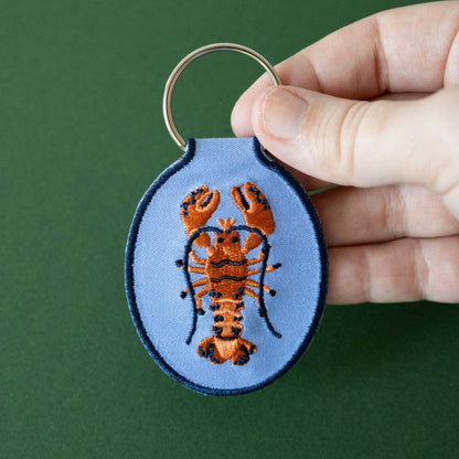 Keychain with embroidered lobster design held by a hand against a green background