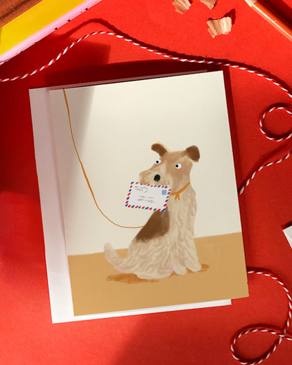 A square greeting card featuring an illustration of a small cream and brown dog holding a postcard with red, white, and blue border details, set against a warm beige ground. The card is displayed on a bright red background with decorative red-and-white twine.