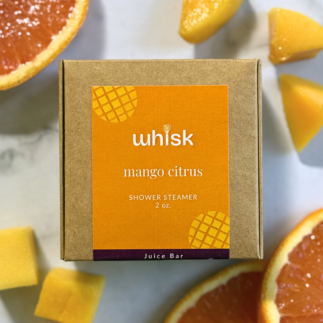 A kraft paper box labeled 'whisk mango citrus Shower Steamer 2 oz.' is centered on a light marble surface, surrounded by sliced grapefruit and mango pieces.