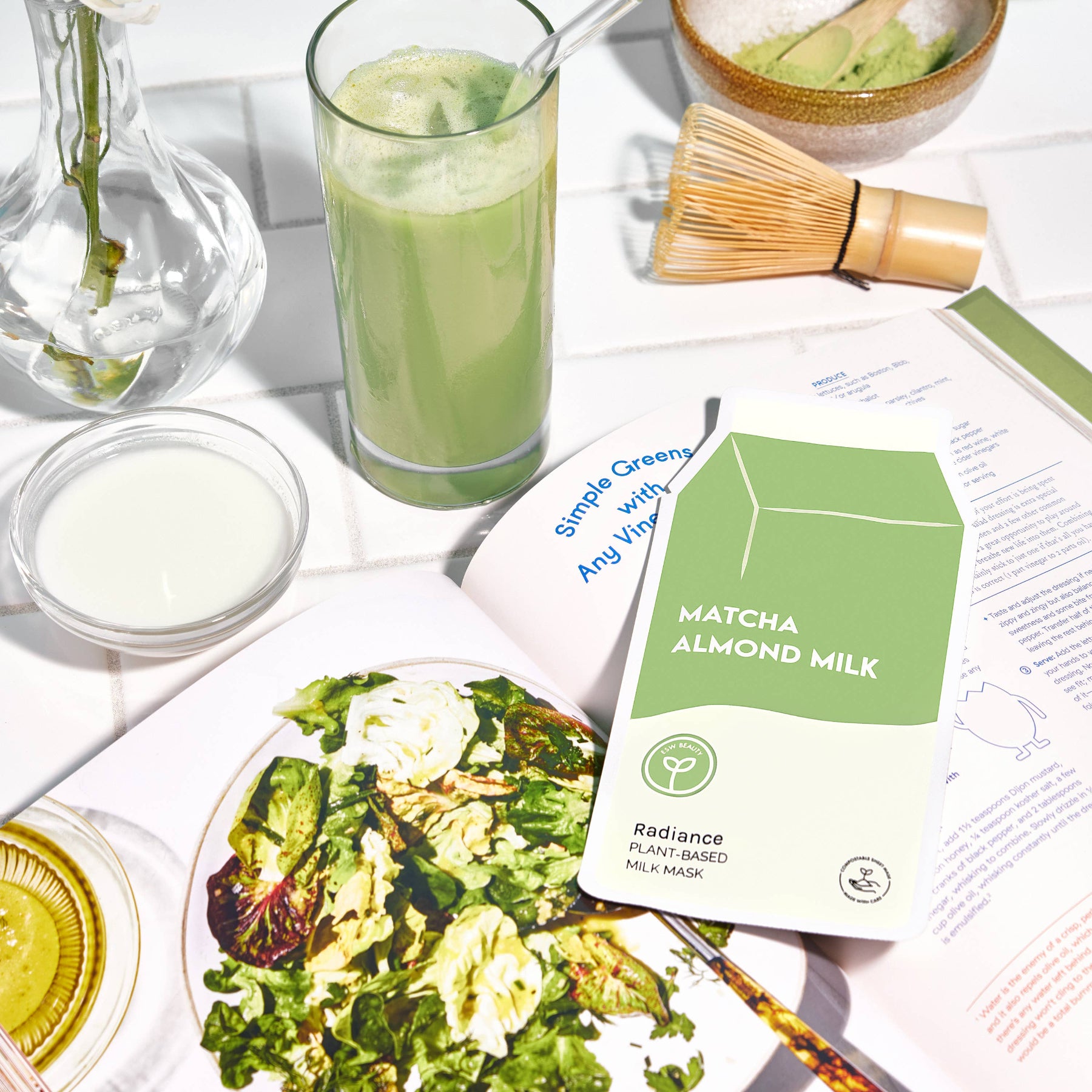 Matcha almond milk package with a glass of matcha, salad, and other green-themed items on a white surface.