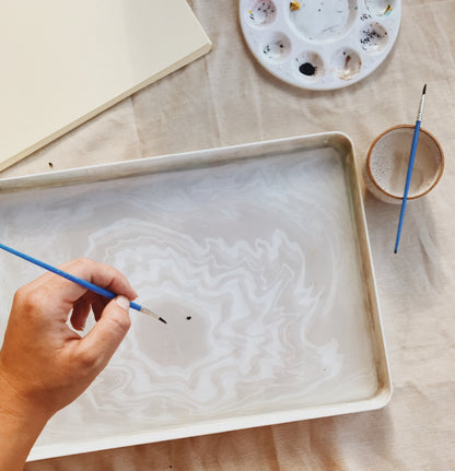 Person painting on a tray with a brush, surrounded by art supplies on a wooden surface.