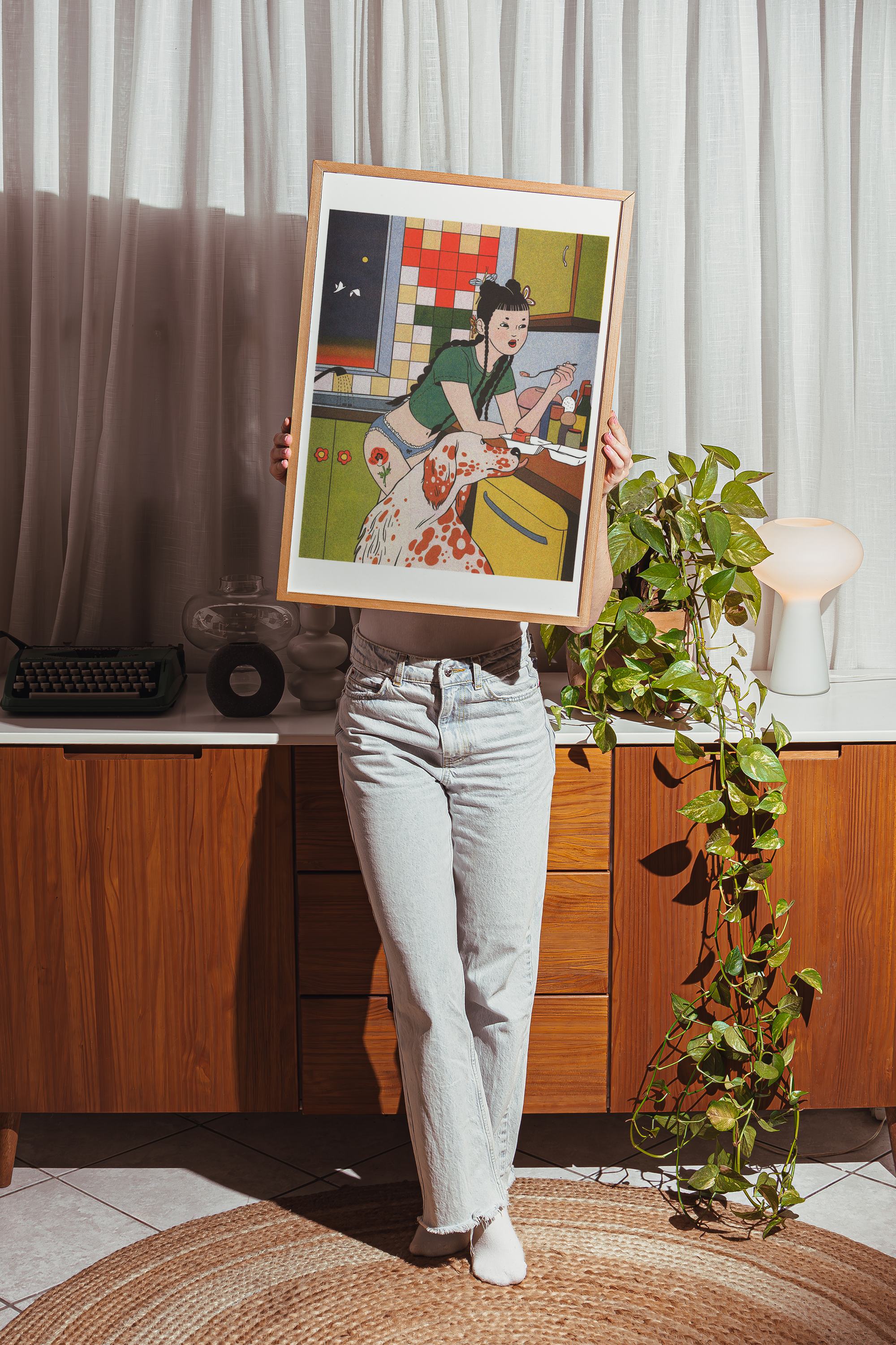 Person holding a framed artwork in a room with wooden cabinets and a plant.