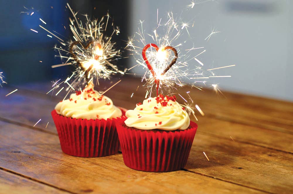 Two cupcakes with red wrappers and white frosting, each topped with a lit sparkler, on a wooden surface.