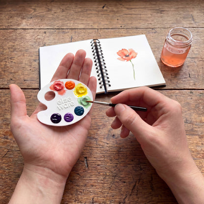 Person holding a paint palette with watercolor paints on a wooden table.
