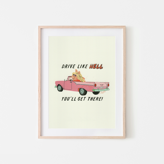 A framed art print featuring a retro pink convertible with MISS PIGGY IN THE DRIVERS seat, with the text 'DRIVE LIKE HELL YOU'LL GET THERE!' on a cream background, set in a light wood frame against a white wall.