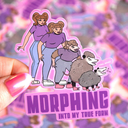 A hand holds a die-cut sticker featuring cartoon characters from the TV show 'Morphing Into My True Form' with purple text reading 'MORPHING INTO MY TRUE FORM'. The sticker has a pink border and is set against a blurred background of other colorful stickers.