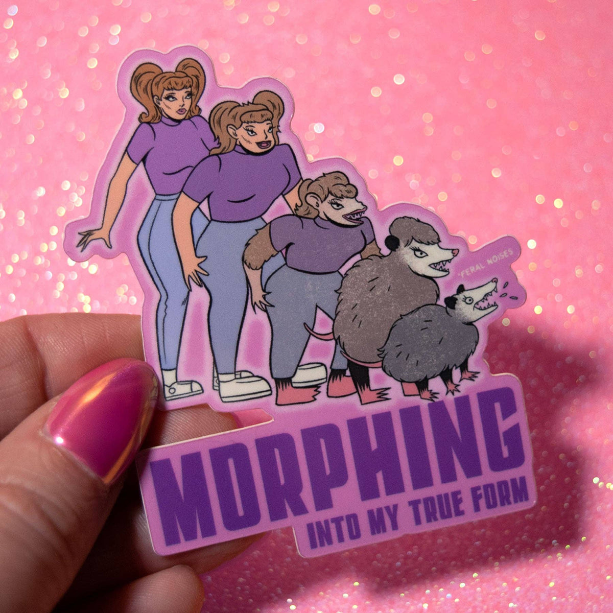 A hand with pink nail polish holds a die-cut sticker featuring two cartoon women and two animals above the text 'MORPHING INTO MY TRUE FORM' on a pink glittery background.