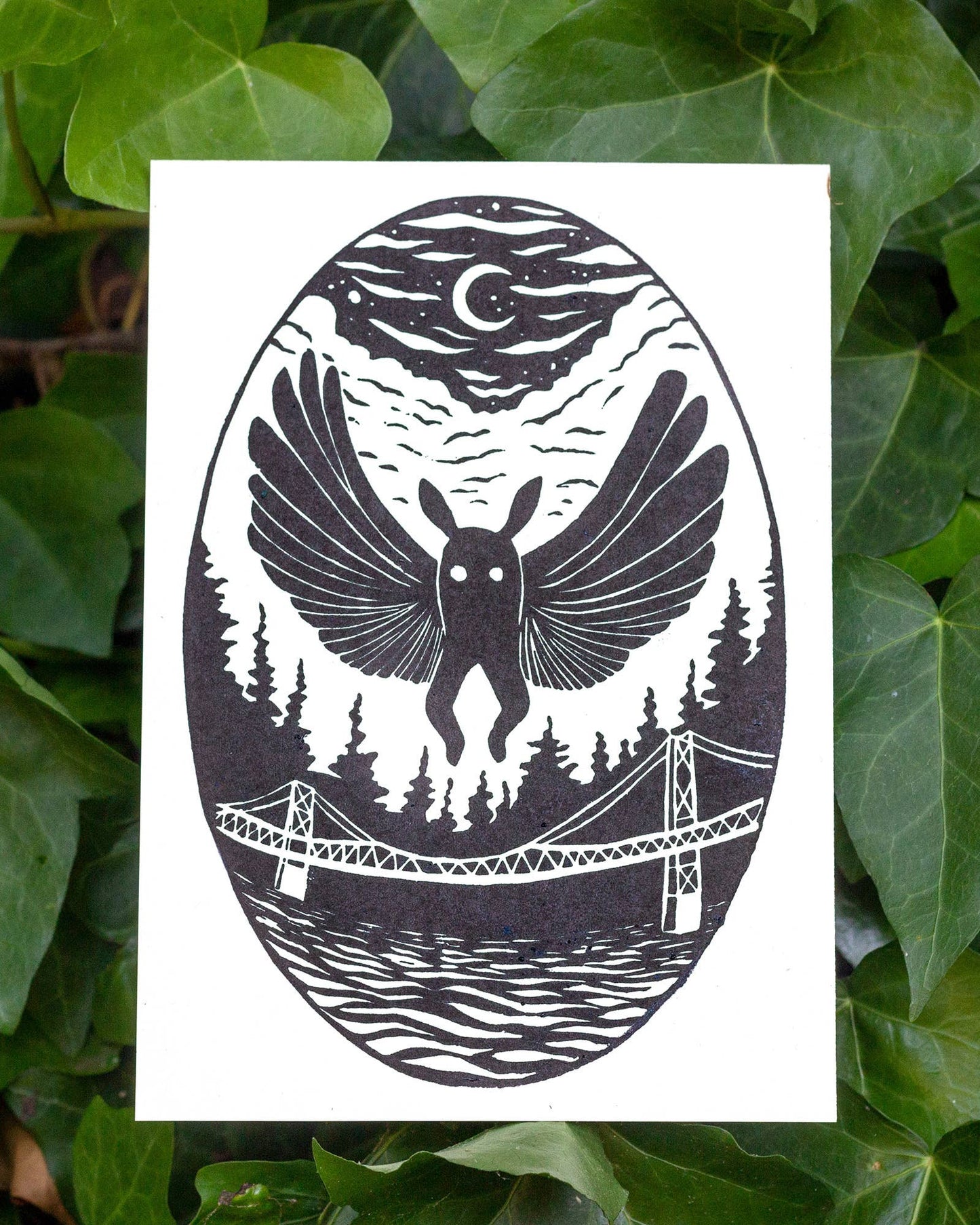 A black and white art print featuring a surreal illustration of mothman over a forest, bridge, and water under a crescent moon, displayed against a background of green ivy leaves.