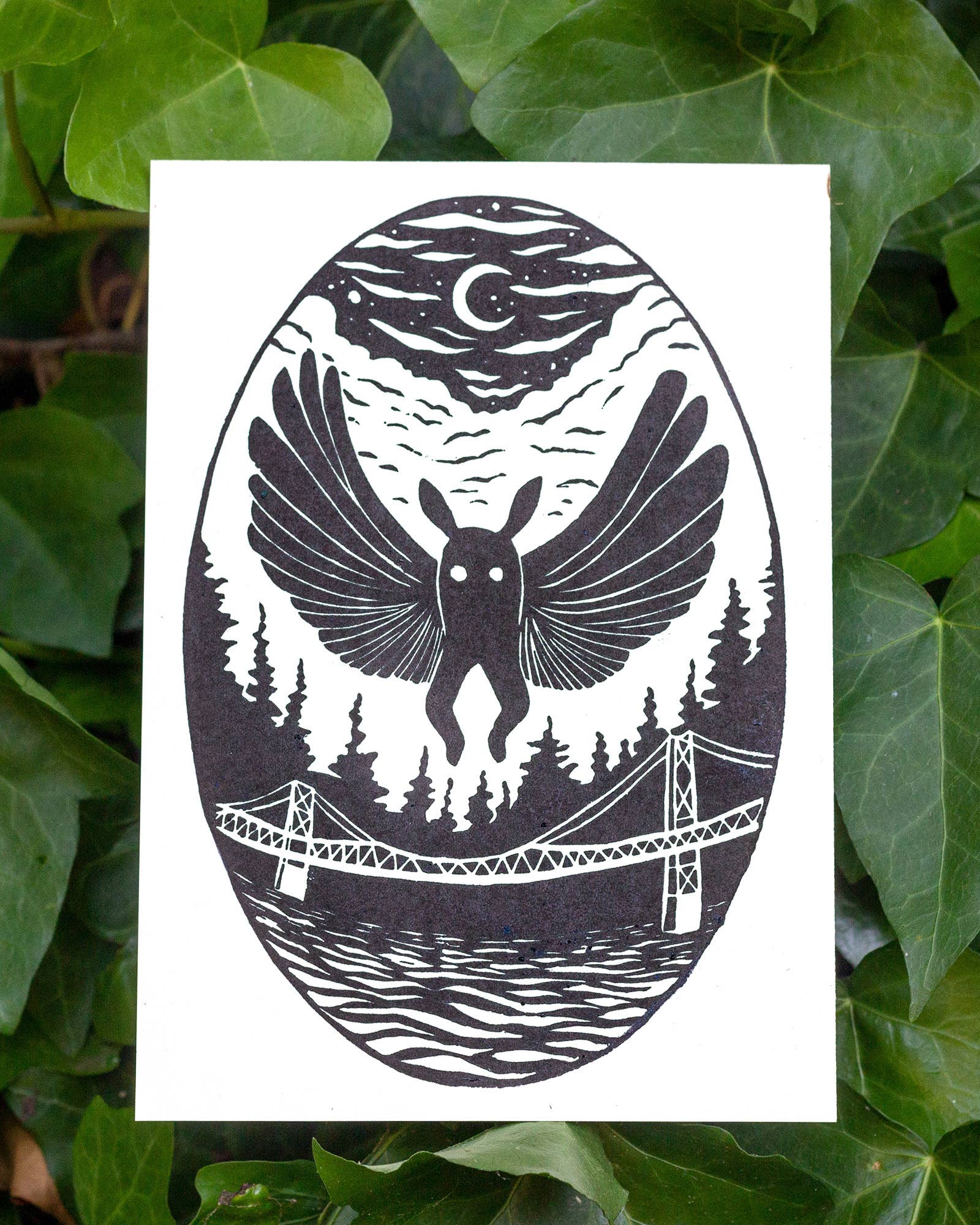 A black and white art print featuring a surreal illustration of mothman over a forest, bridge, and water under a crescent moon, displayed against a background of green ivy leaves.