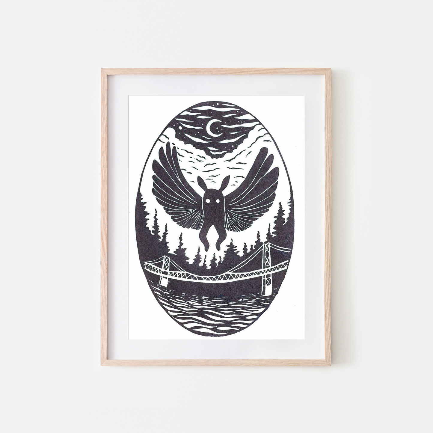 A framed art print featuring a black and white linocut-style illustration of a bird with spread wings over a forest, bridge, and water, under a crescent moon, shown in a light wood frame against a white wall.