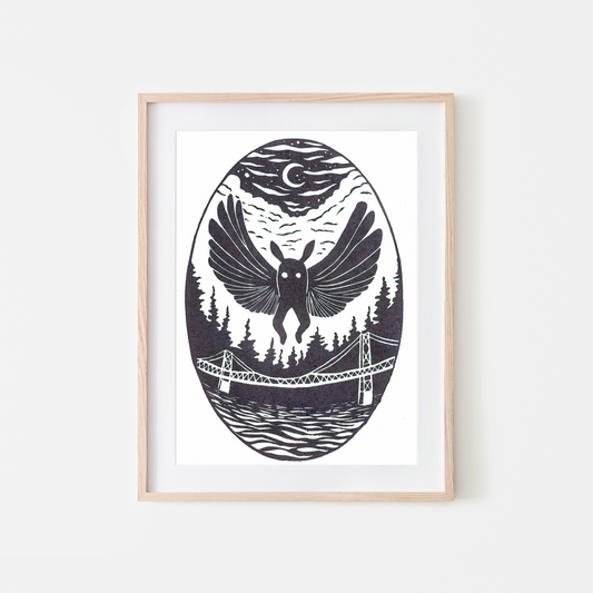 A framed art print featuring a black and white linocut-style illustration of a bird with spread wings over a forest, bridge, and water, under a crescent moon, shown in a light wood frame against a white wall.