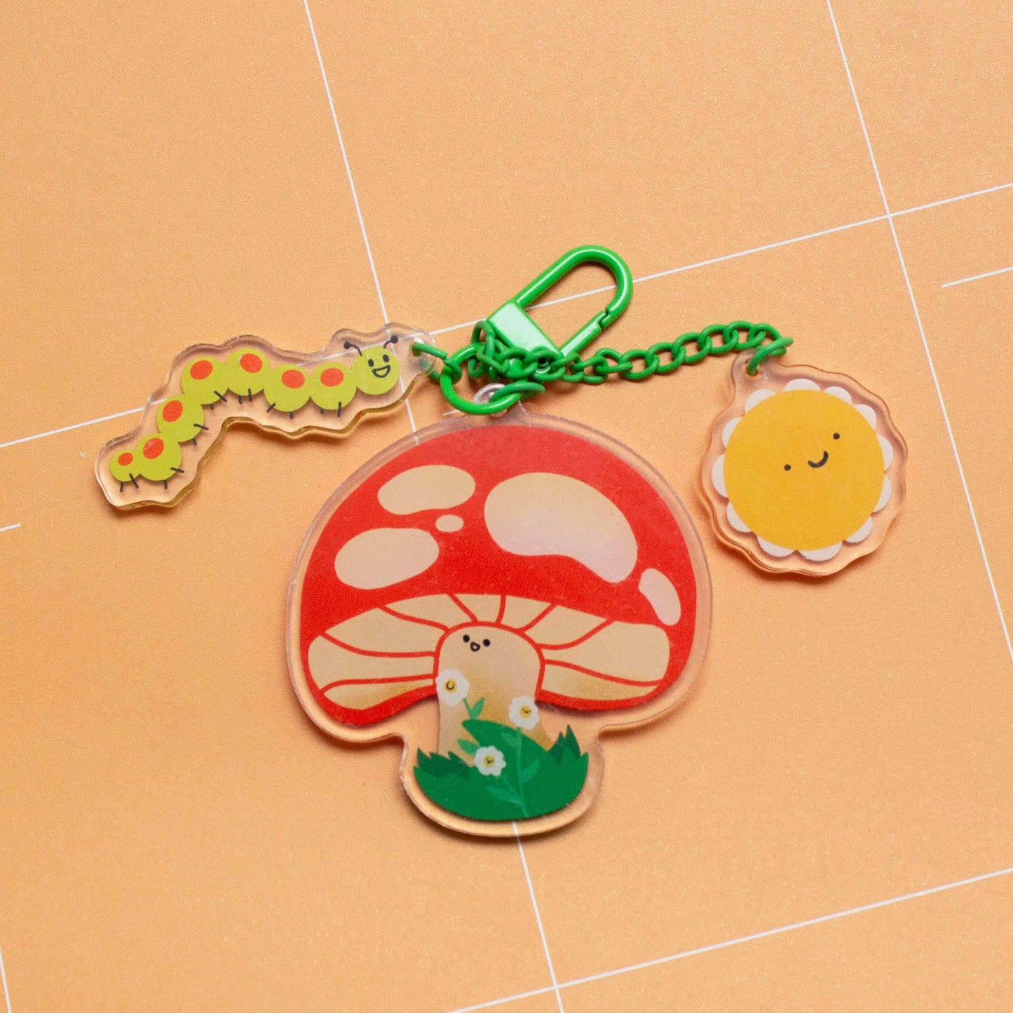 A cute acrylic keychain featuring a red and white mushroom with a small white bunny, a green caterpillar, and a yellow sun on a green chain with a silver clasp, set against a peach-colored grid background.