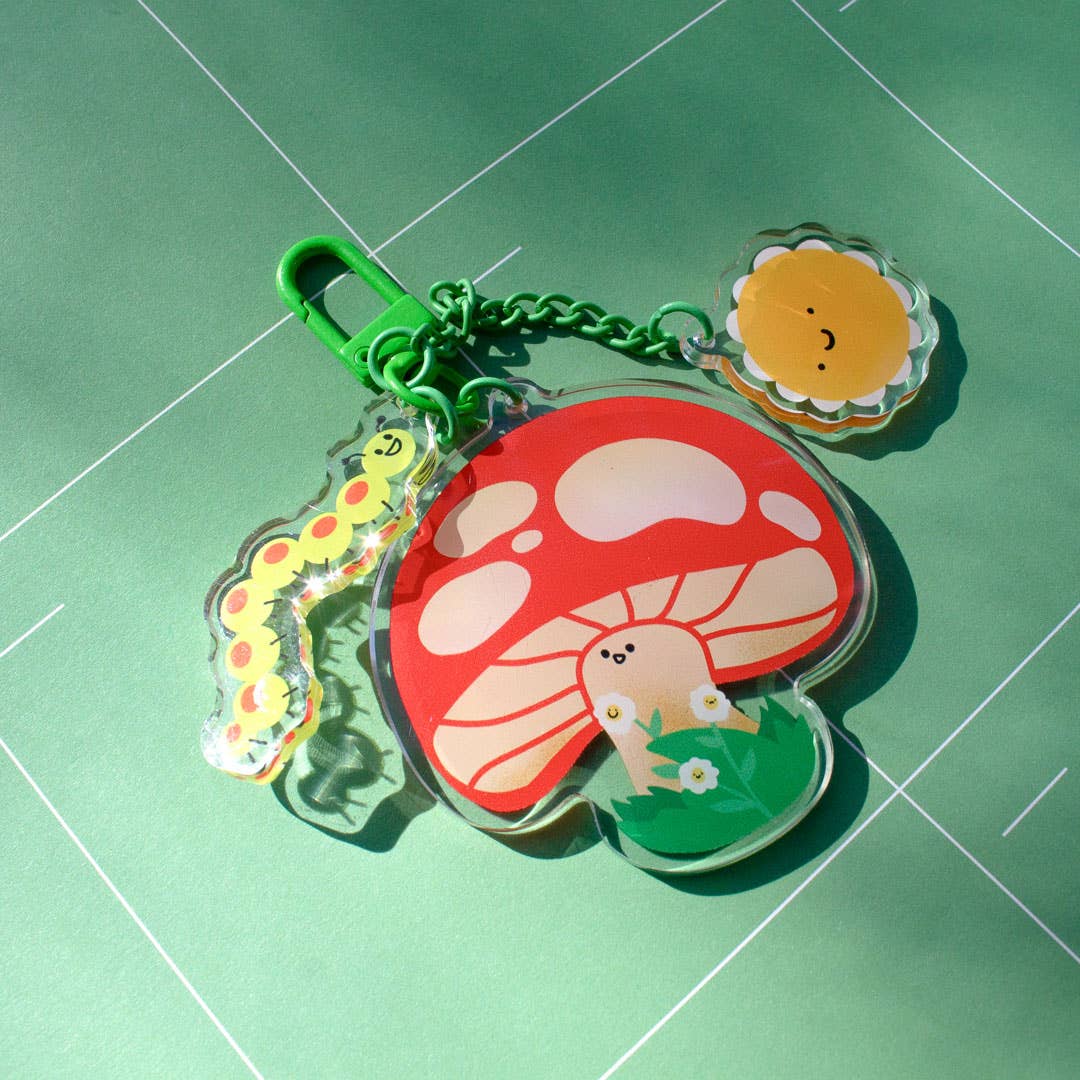 A colorful acrylic keychain featuring a red and white mushroom, a yellow caterpillar, a smiling sun, and a small character in a green field, attached to a green clasp on a green background.