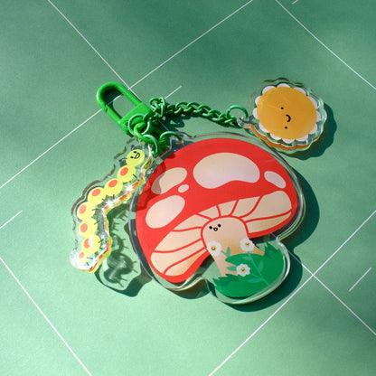 A colorful acrylic keychain featuring a red and white mushroom, a yellow caterpillar, a smiling sun, and a small character in a green field, attached to a green clasp on a green background.