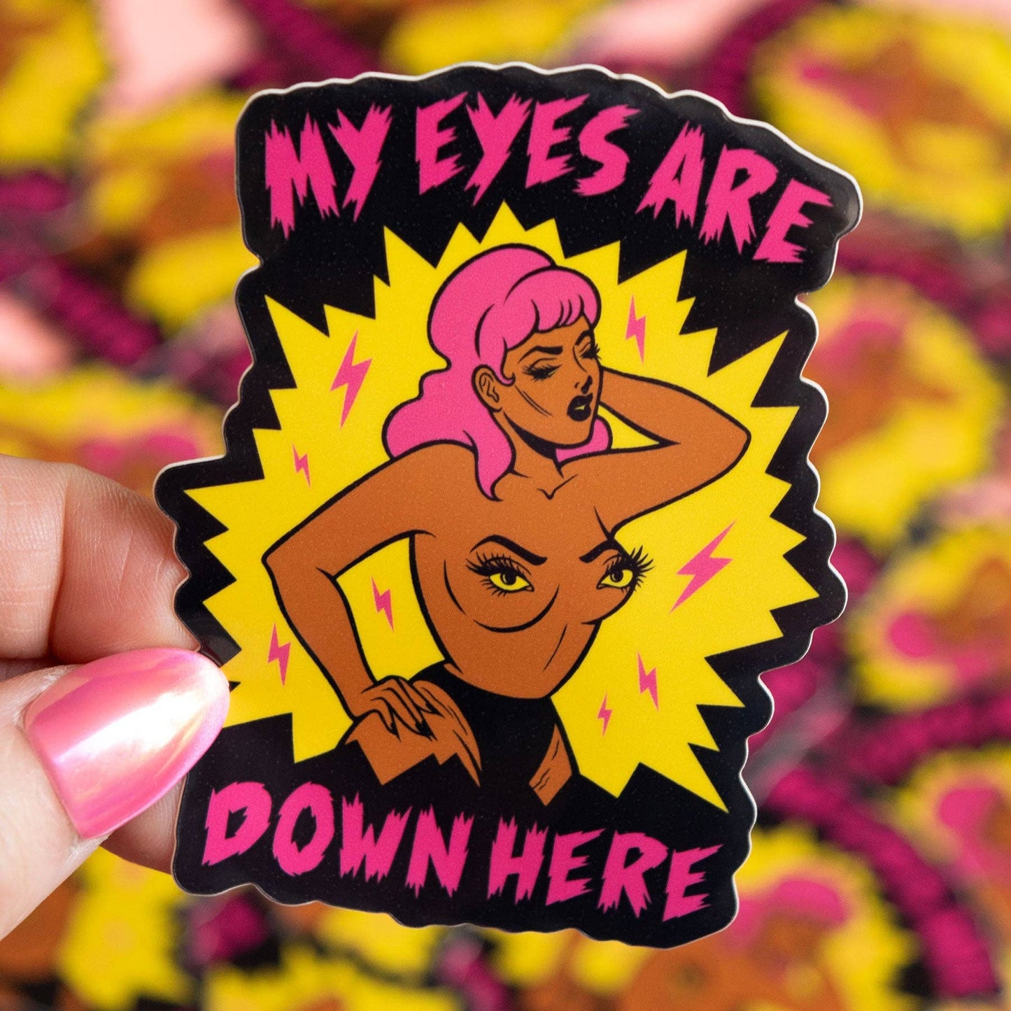 A hand holds a die-cut sticker featuring a retro-style illustration of a woman with pink hair and the text 'MY EYES ARE DOWN HERE' on a black background with a yellow starburst.