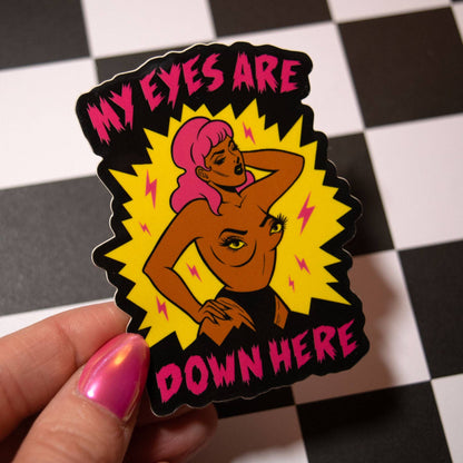 A hand with pink nail polish holds a die-cut sticker featuring a stylized illustration of a woman with pink hair and the text 'MY EYES ARE DOWN HERE' on a black background, set against a black-and-white checkered floor.